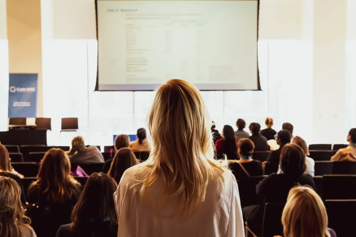 A woman seen from behind at the back of a bright conference hall, facing rows of seated attendees and a large presentation screen.