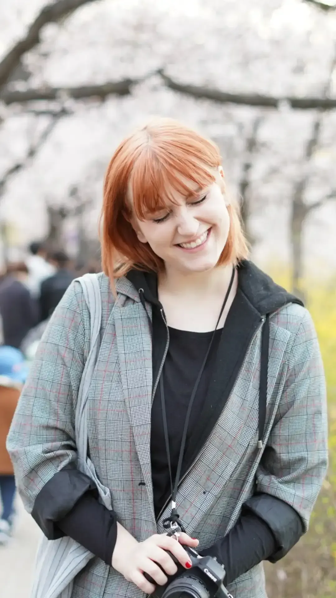 Jane Hargrave laughing while holding a camera, wearing a plaid blazer and a black hoodie in a park with cherry blossoms in the background