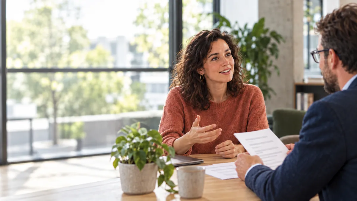Een vrouwelijke kandidaat in een roestkleurige trui praat aan een houten tafel met een recruiter die een cv vasthoudt. Ze zitten in een lichte kantoorruimte met grote ramen en planten.