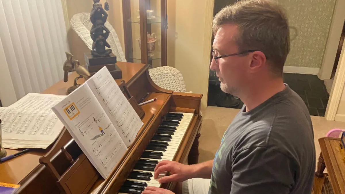 An adult, dressed in a colorful long sleeve and eyeglasses, sits attentively at a sleek digital keyboard, their fingers moving gracefully over the keys. The soft glow of a tilt light bathes the scene, casting an inviting, warm ambiance around them. 