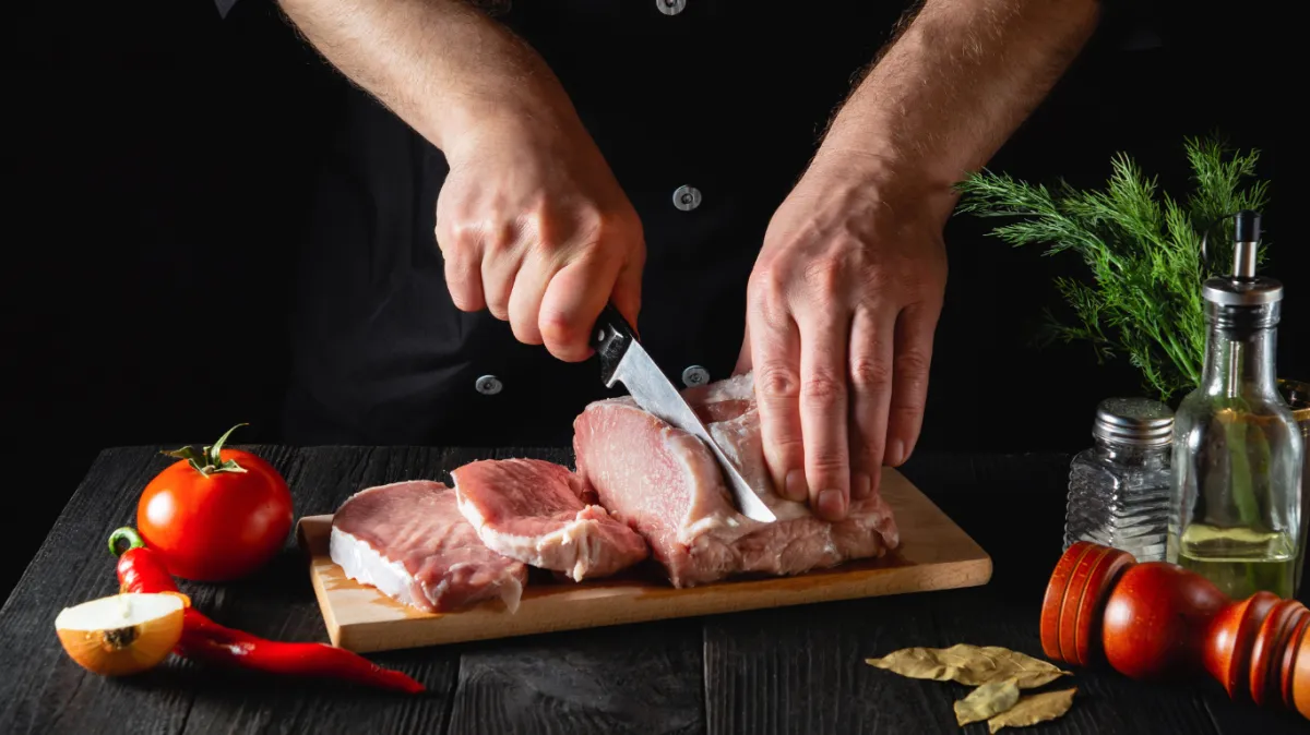 Person in black slicing a slab of organic, grass-fed beef on a wooden table, surrounded by fresh tomatoes, vegetables, herbs, salt, and oil, highlighting farm-to-table quality from Briarmead Farm.