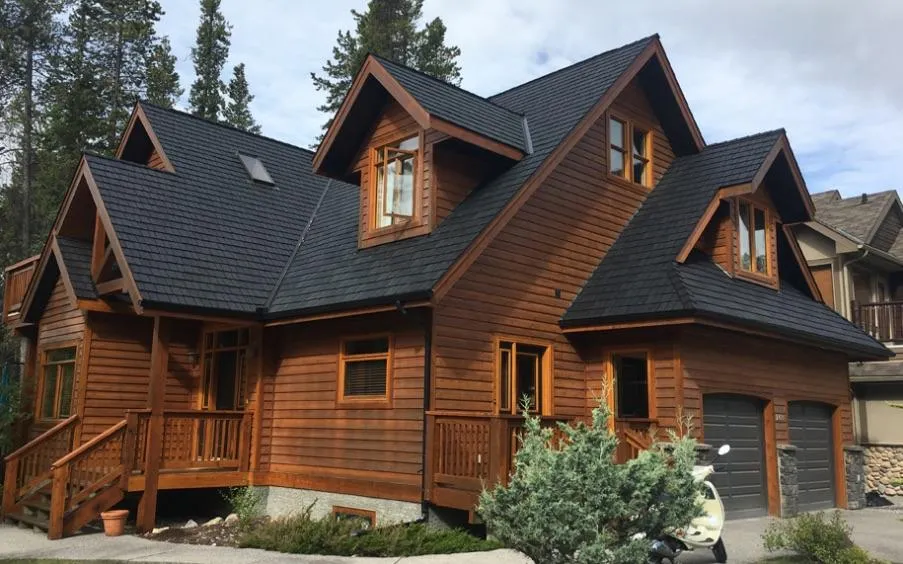 A two-story wooden house with gabled roofs, a front porch, and an attached double garage, surrounded by trees and greenery.