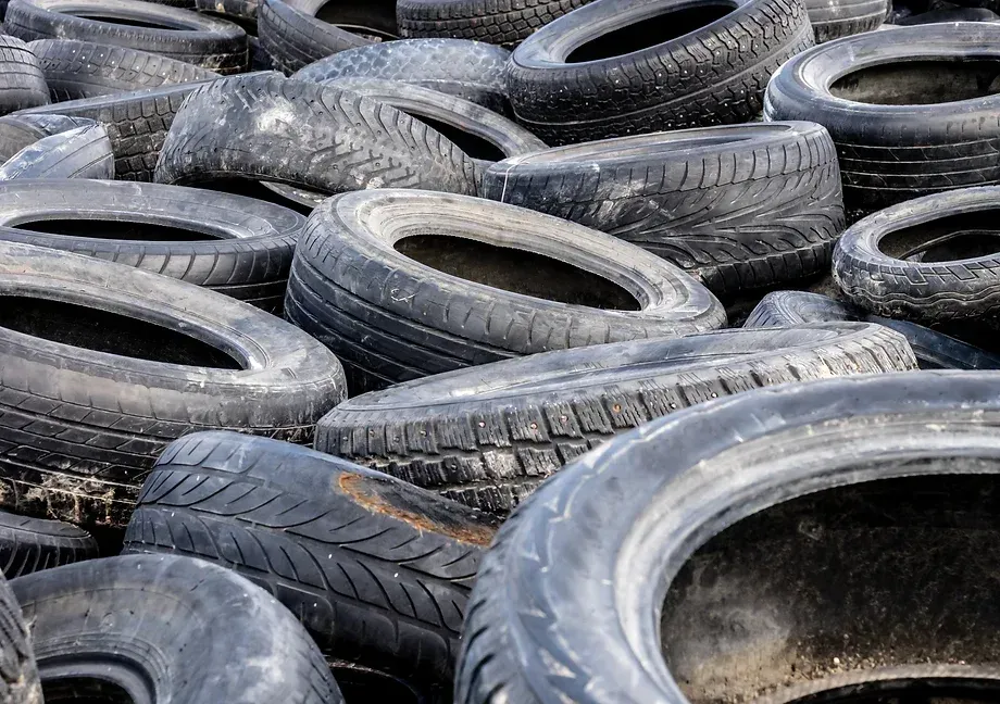 A large pile of used tires stacked haphazardly in an outdoor setting. The tires vary in size and condition, showing signs of wear and tear with some dirt and debris visible. The image conveys a sense of clutter and environmental waste.