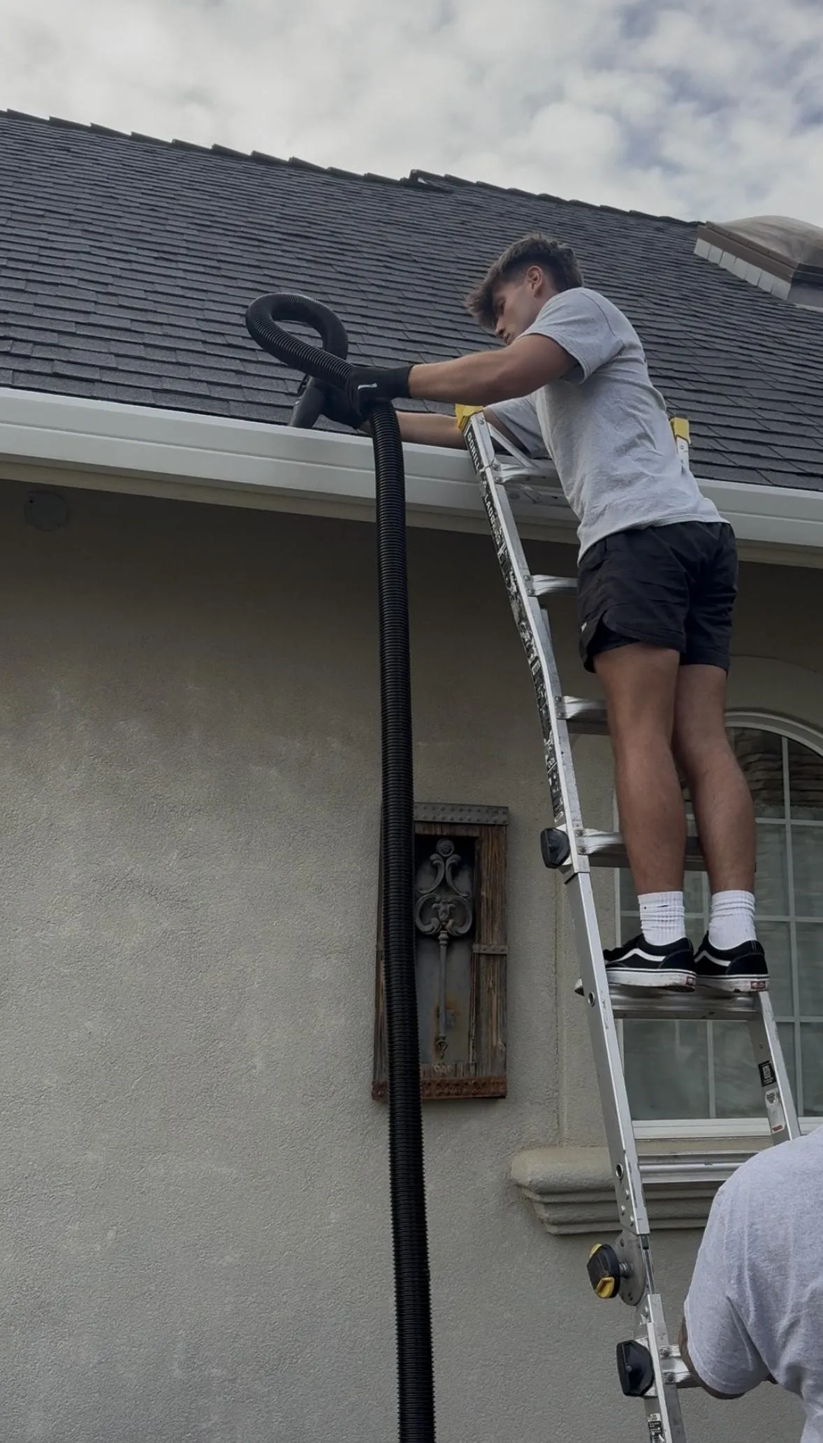 Technician cleaning a home gutter, removing leaves with safety gear; clear view of gutter maintenance.
