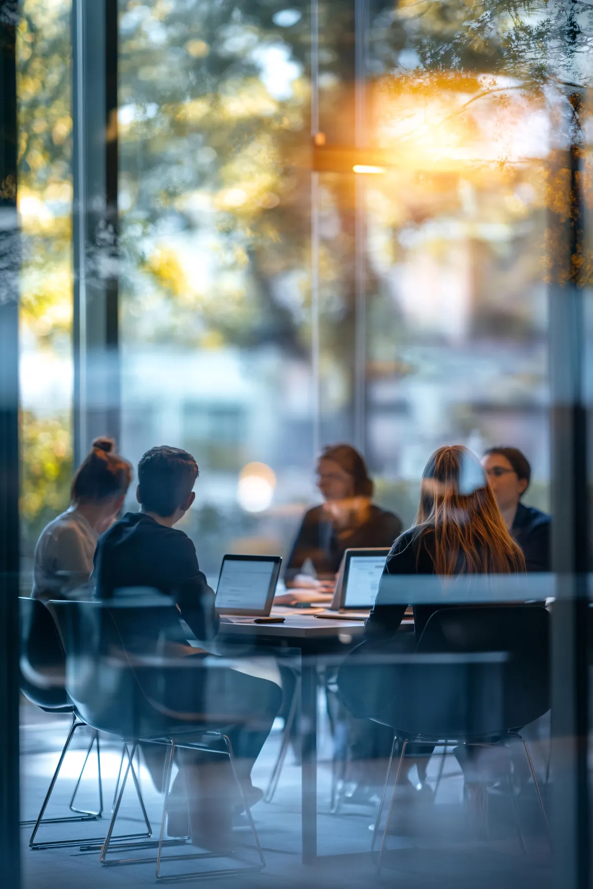 Team meeting in a modern glass-walled office with professionals seated around a table using laptops and collaborating in a corporate workspace.