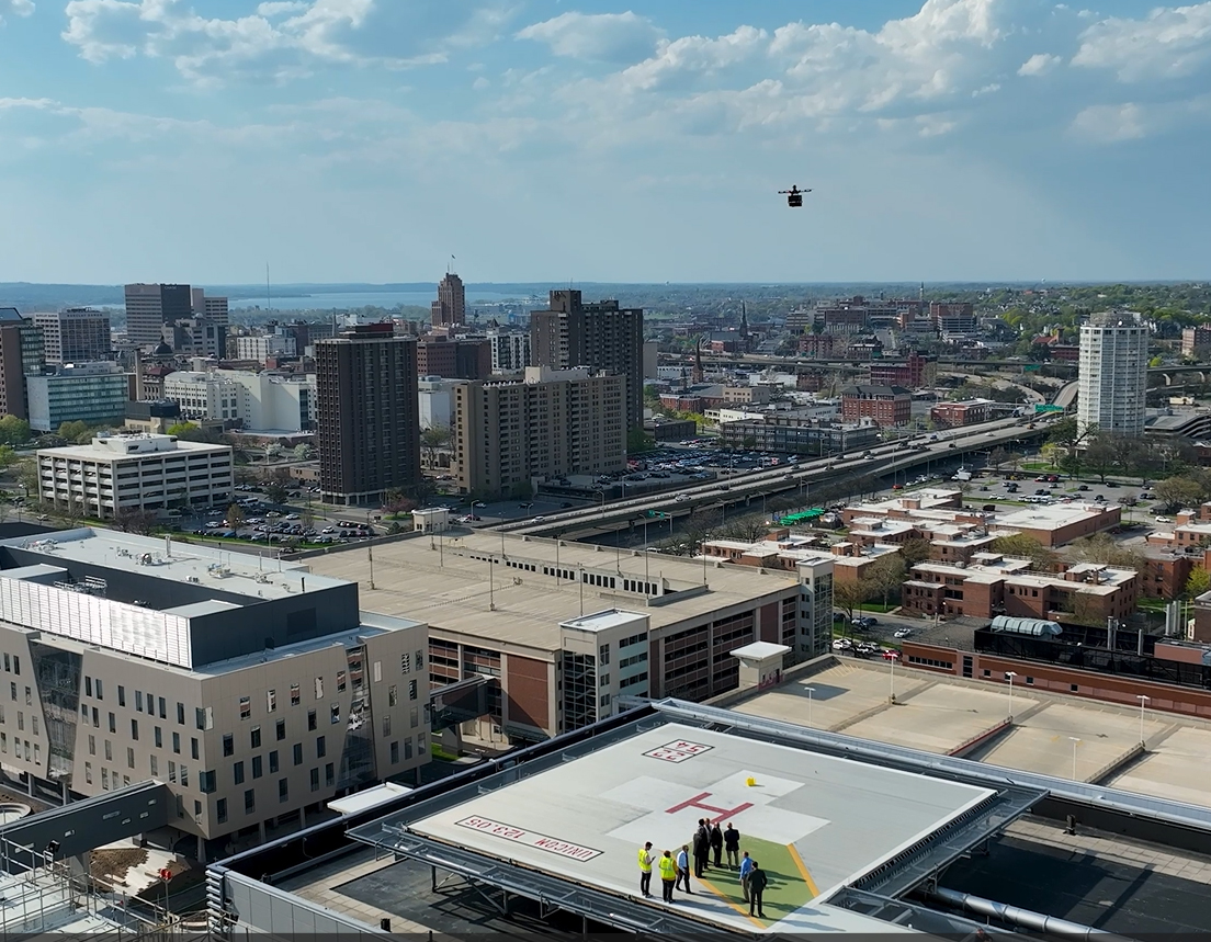 Aerial drone view of a city skyline with a rooftop hospital helipad, safety personnel in high-visibility vests, and a helicopter approaching above urban buildings under a blue sky.
