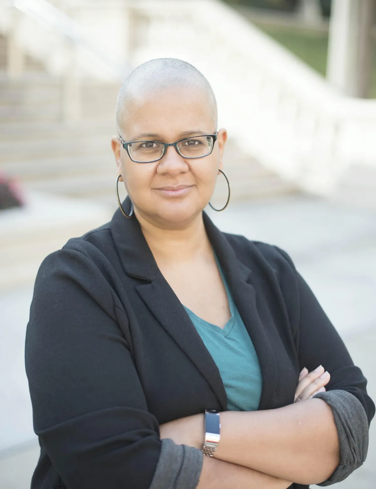 A picture of 'Tine Zekis in glasses, and hoop earrings wearing a blazer and green shirt, standing with arms crossed.