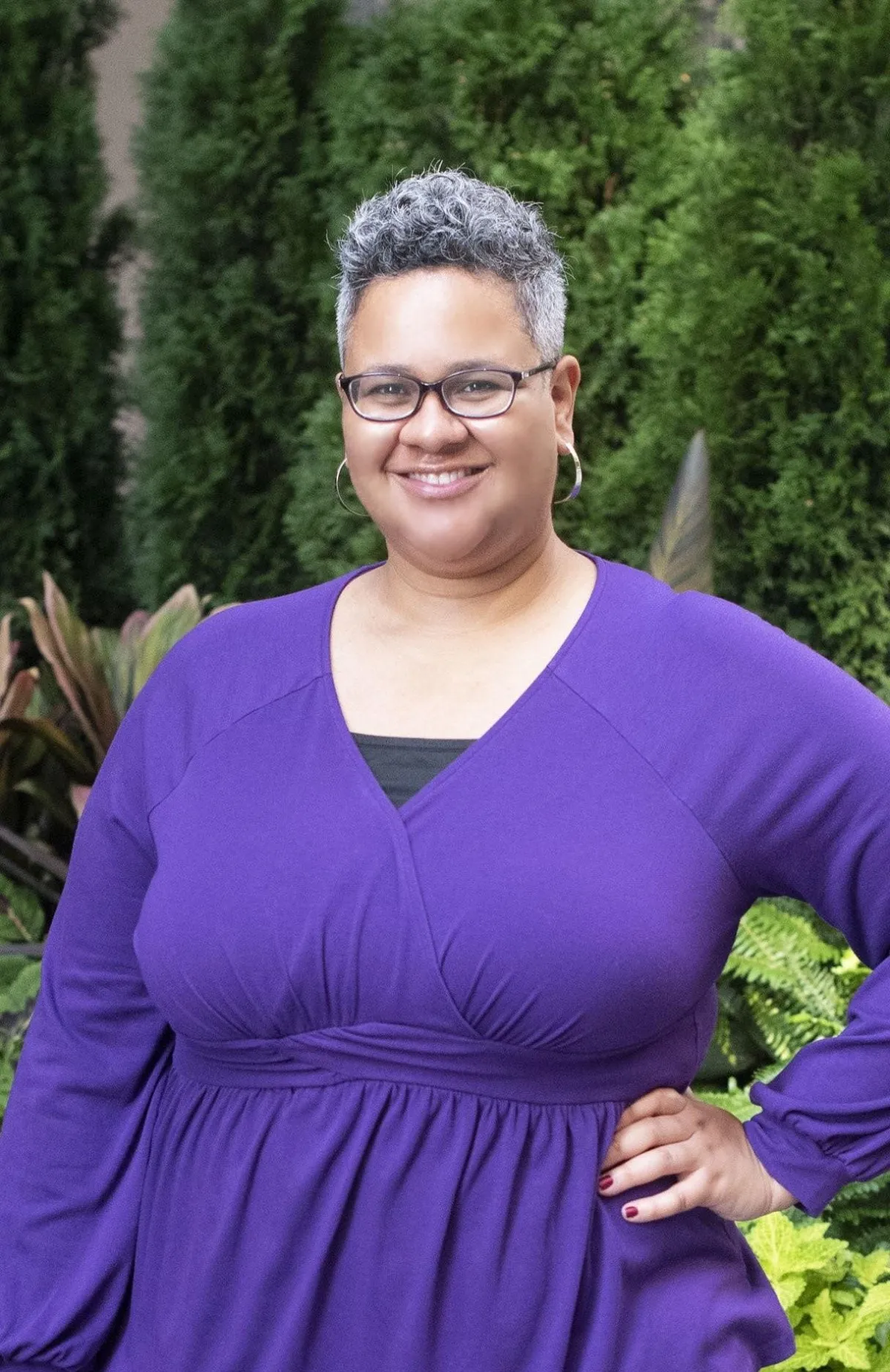 Tine Zekis smiling with short gray hair and glasses, in a purple top, standing before lush greenery.