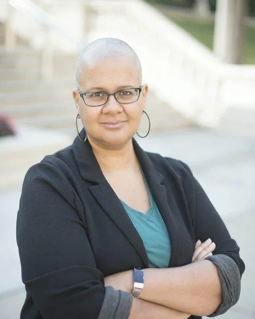 A picture of 'Tine Zekis in glasses, and hoop earrings wearing a blazer and green shirt, standing with arms crossed.
