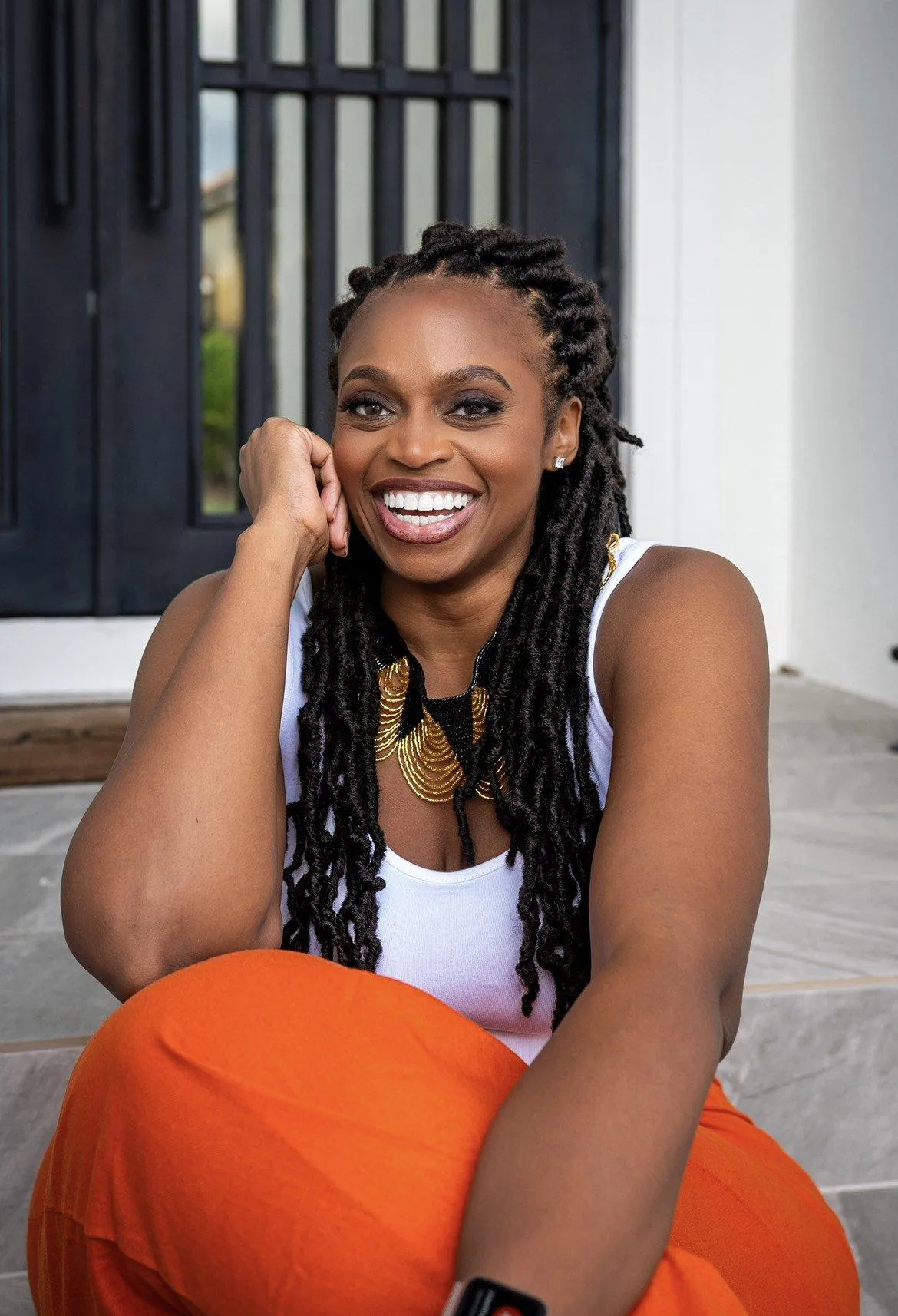 Ileka Falette smiling with long locs, wearing a white top and orange pants, seated on a doorstep.