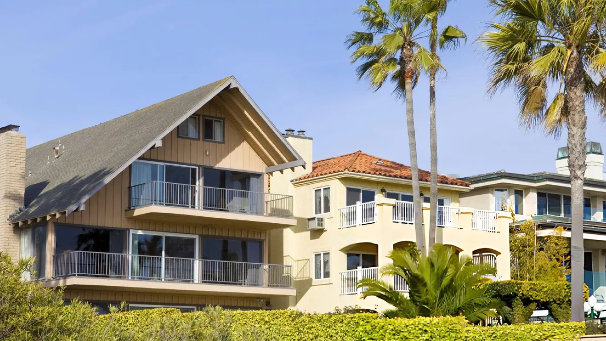 Sunny coastal neighborhood with a wooden gabled house, a yellow tile-roofed home, and tall palm trees against blue sky.