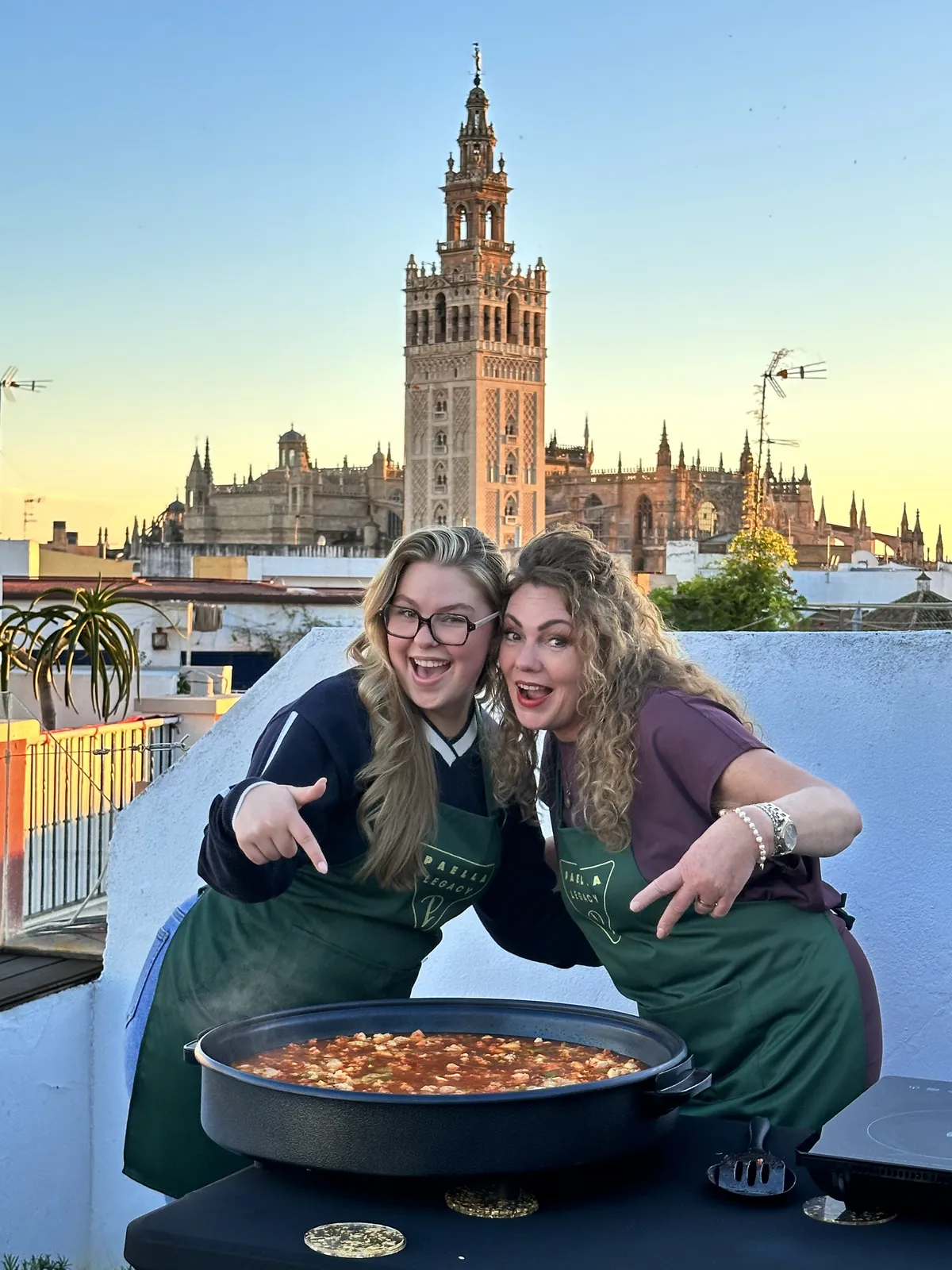 Four guests in Paella Legacy aprons laughing beside their finished paella at sunset, the Giralda glowing behind them