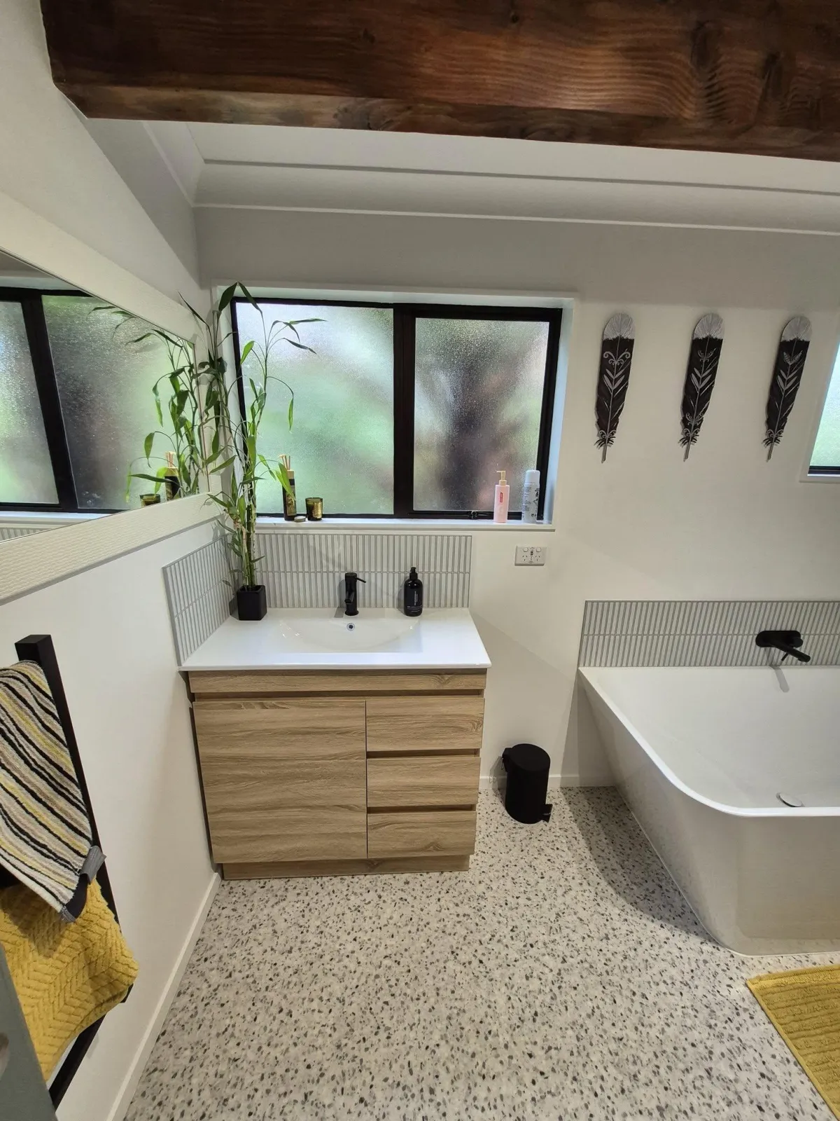 Stylish bathroom featuring a timber vanity, freestanding tub, and frosted windows, with indoor plants and Māori-inspired wall art.
