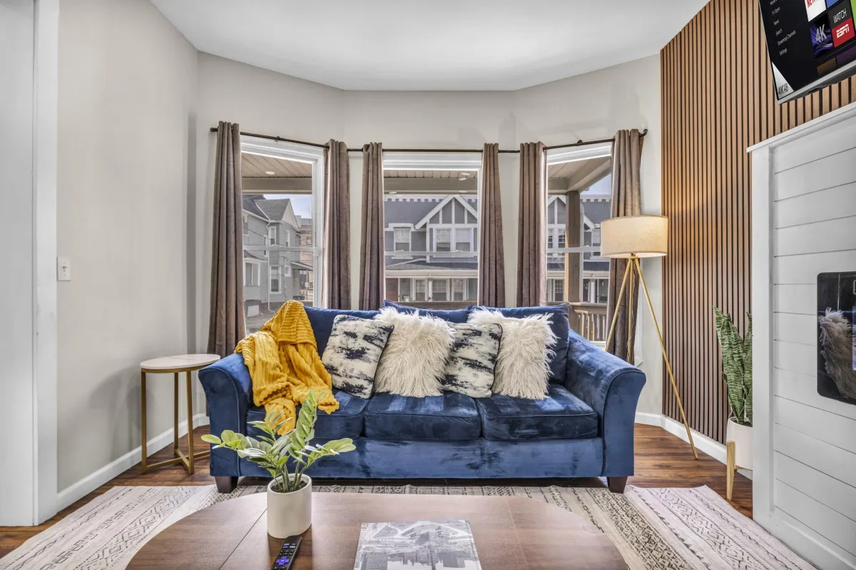 Bright short-term rental living room with a blue velvet sofa, throw pillows, and large windows
