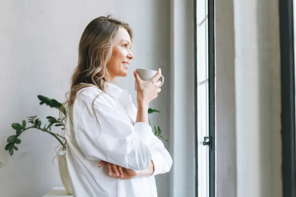 Woman in her fifties by a sunlit window holding a warm drink, looking calm and at ease in midlife