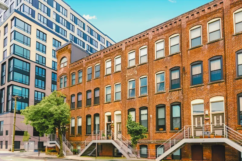 A row of historic red brick townhouses with tall arched windows and raised front stoops, set along a quiet street. Behind them rises a modern multi-story apartment building with glass and white paneling, creating a contrast between old industrial-style architecture and contemporary design. A leafy tree stands near the sidewalk in front of the buildings under a clear blue sky.