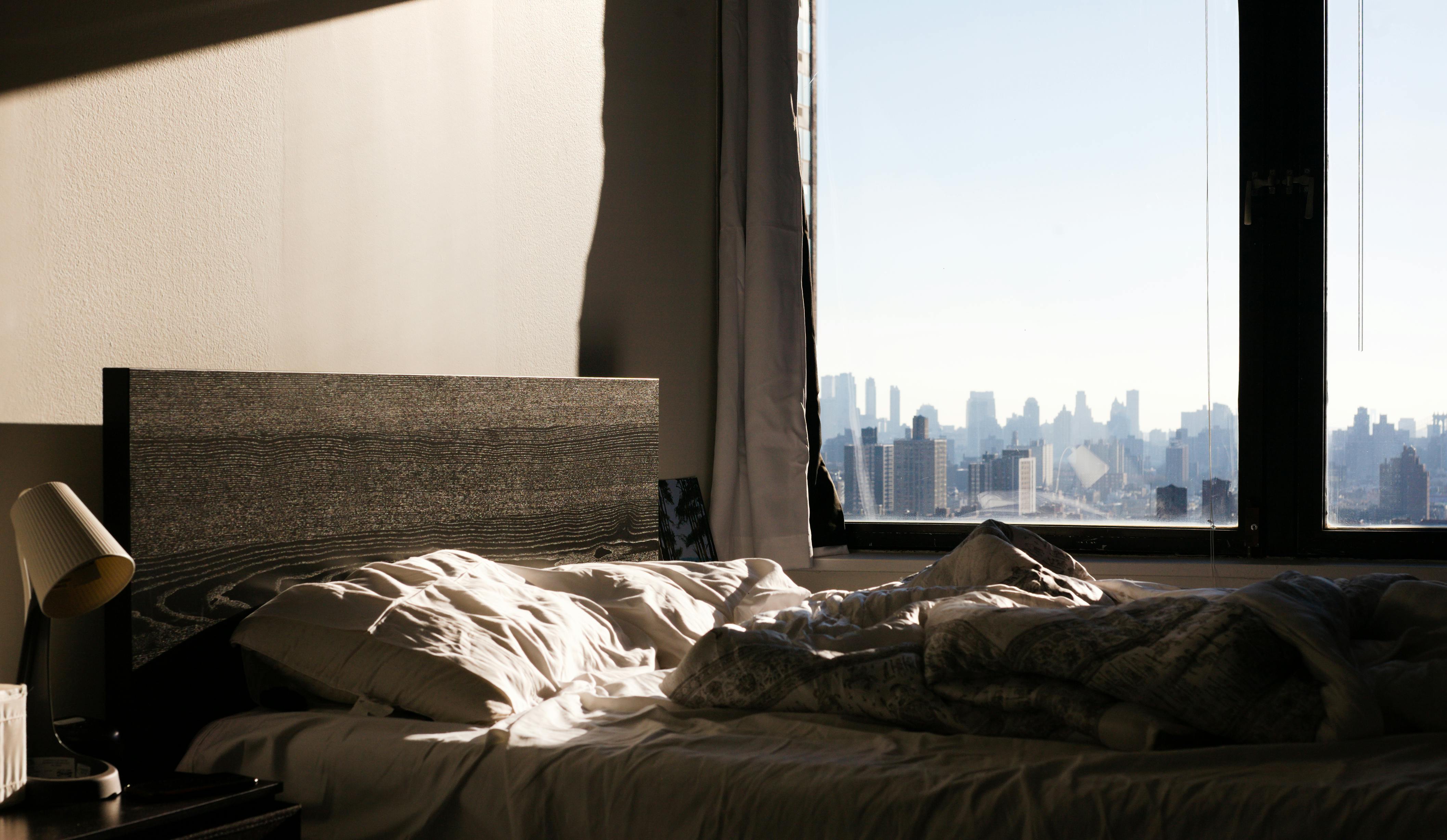Sunlit modern bedroom overlooking the Manhattan skyline with unmade bed and warm morning light streaming through large windows.