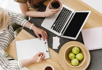 Two women with unpredictable energy building business together. Top down view- hands, computer, notepad, coffee cup. 