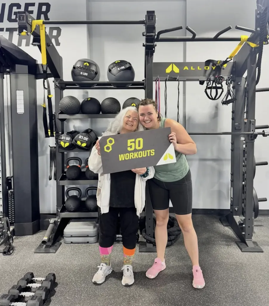 Two Alloy Personal Training members pose for a photo, smiling and holding a sign that reads “50 Workouts.” The gym background features strength training equipment. The image highlights small group training, personalized workout plans, and functional strength training for adults 35+ at Alloy Personal Training.