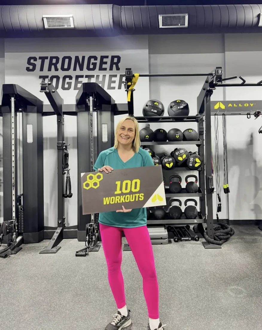 A smiling Alloy Personal Training member proudly holds a sign showing "100 Workouts" in front of gym equipment, with the "Stronger Together" slogan visible in the background. The gym focuses on strength training for weight loss, personalized workout plans, and functional strength training for adults 35+.