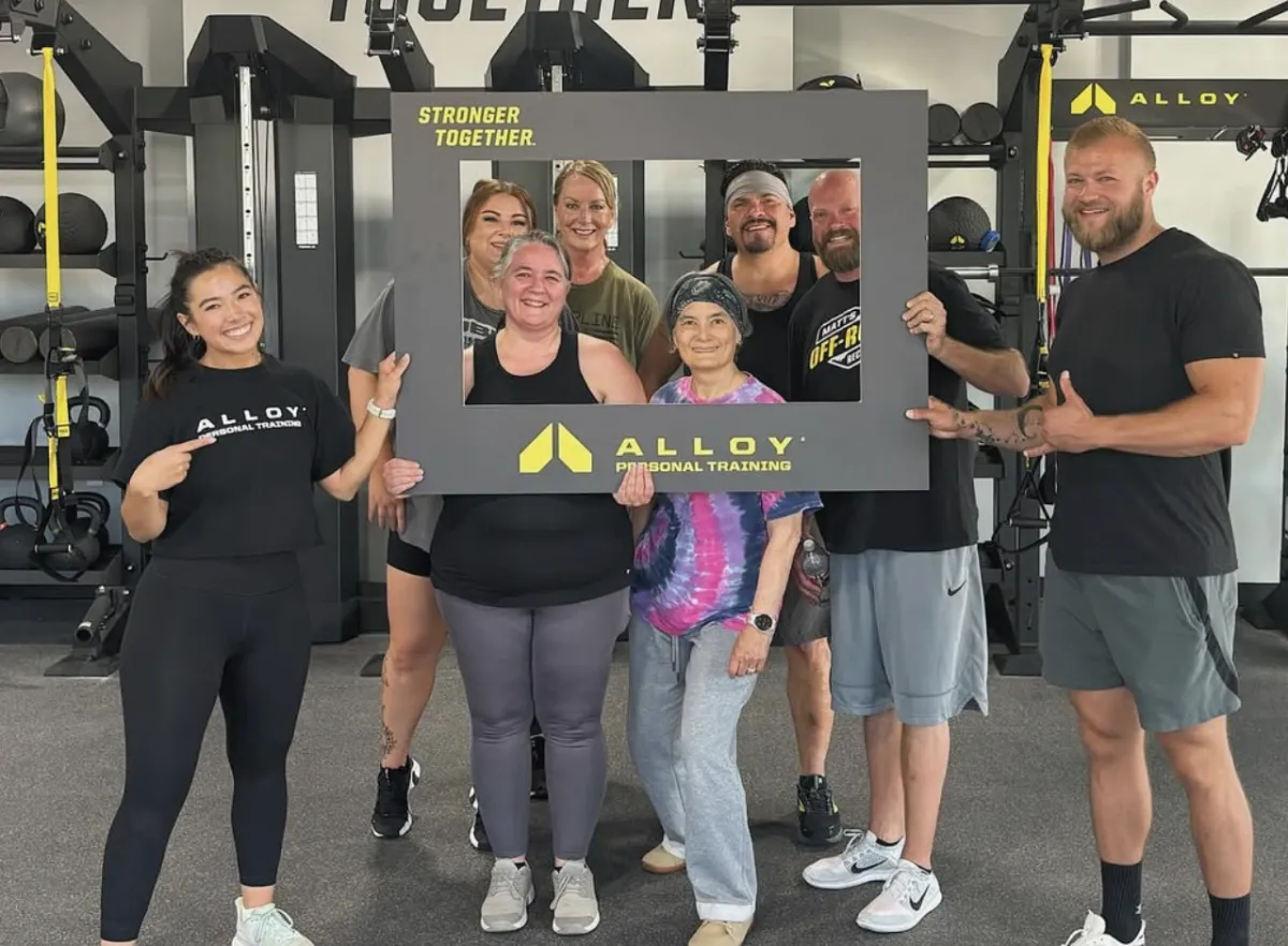 A group of Alloy Personal Training members smiles as they pose with a “Stronger Together” sign after a workout session. The group is diverse, with individuals of different ages and fitness levels, emphasizing the supportive community at Alloy. In the background, fitness equipment is visible, showcasing the gym’s focus on strength training, functional strength training, and personalized fitness programs for all.