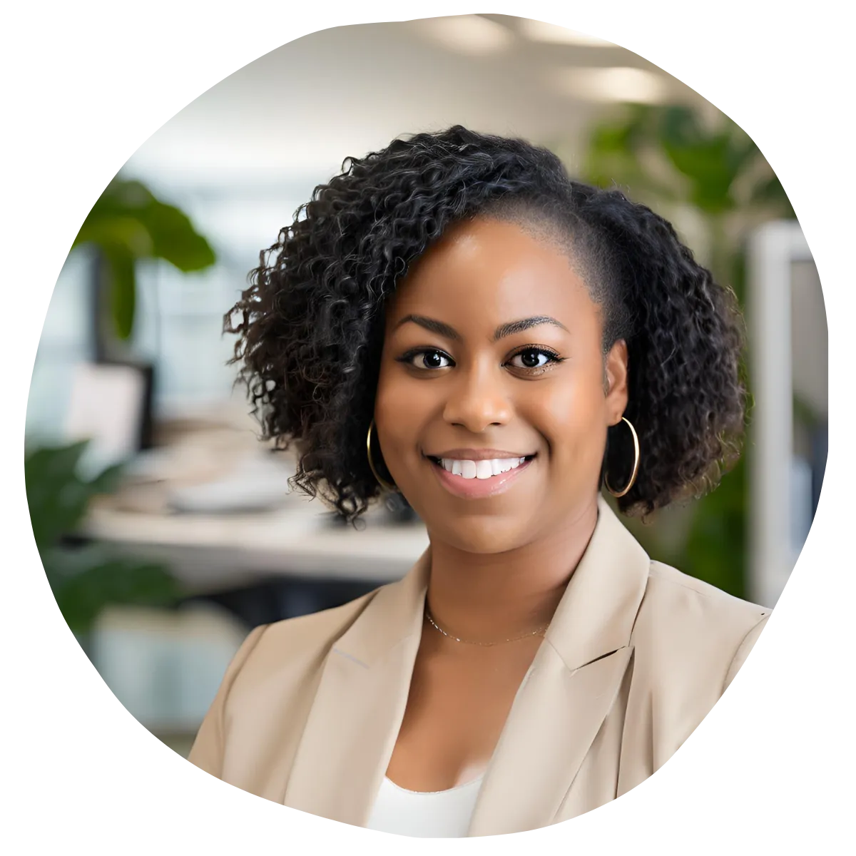 A woman with curly hair, wearing a beige blazer and hoop earrings, smiles at the camera. She is in an office setting with blurred background elements such as desks and plants.