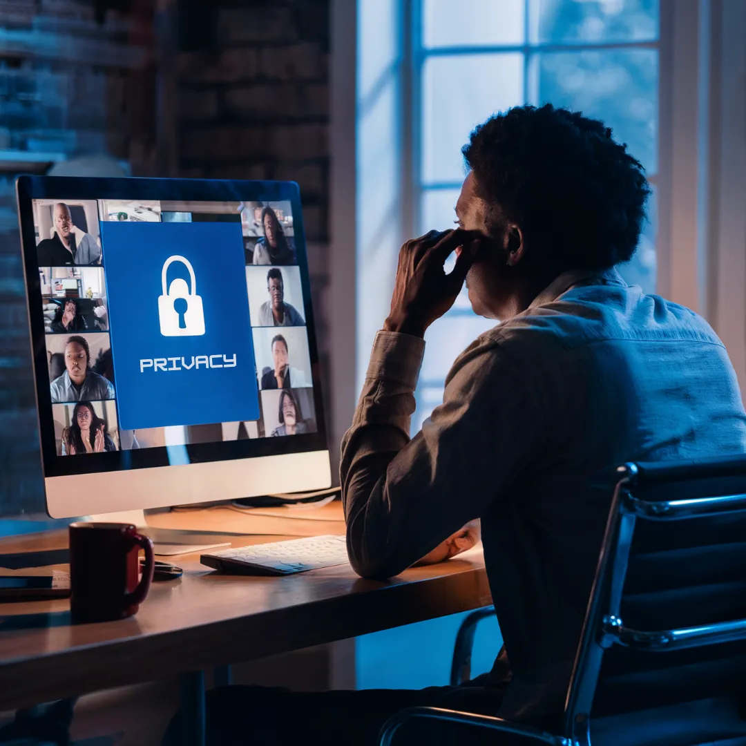 A person sits at a desk with a large computer screen displaying a video conference call with multiple participants. The center of the screen shows a padlock icon with the word "PRIVACY" beneath it. The setting suggests a home office with dim, blue-toned lighting.