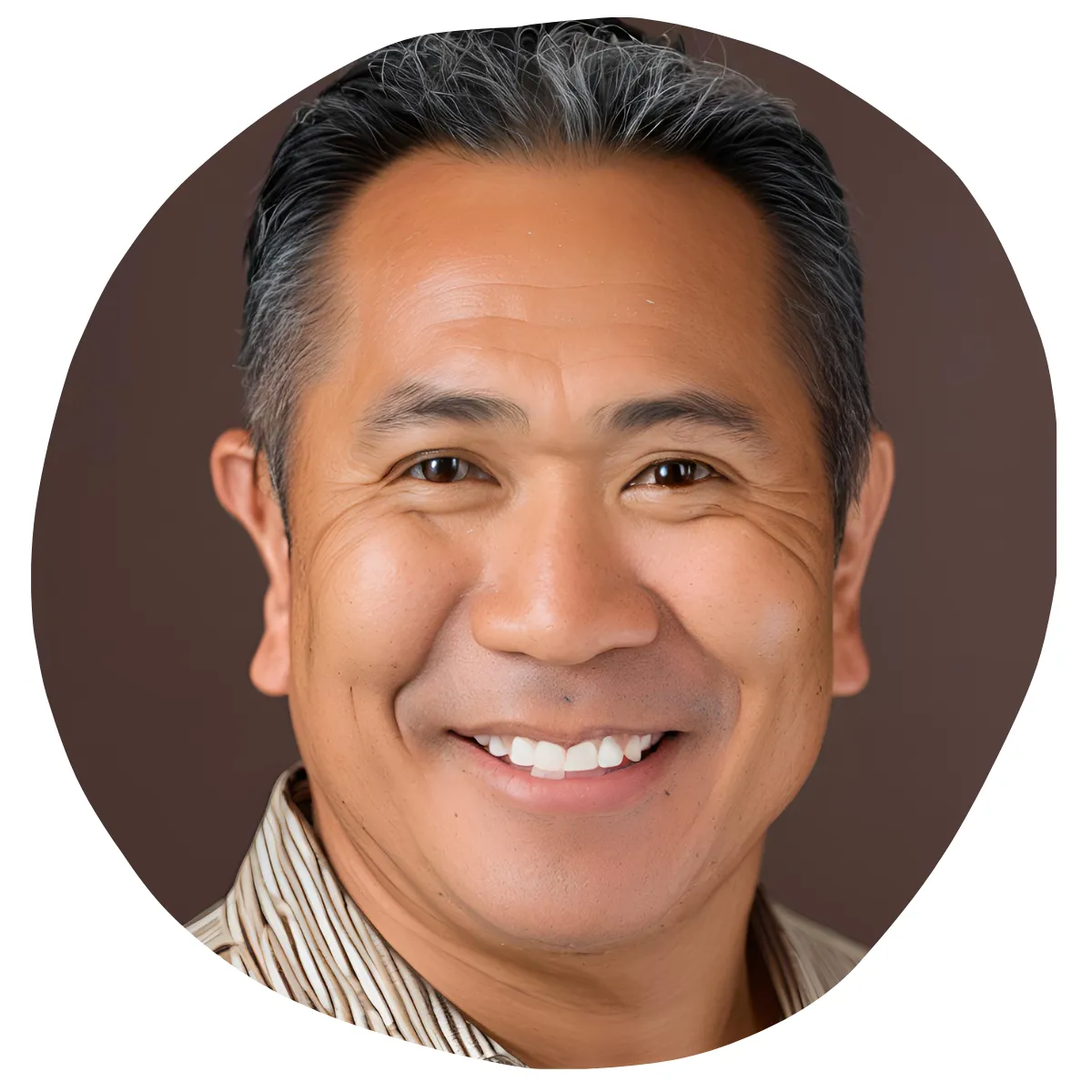 A man with short black and gray hair smiles warmly at the camera. He has a smooth skin complexion with visible laugh lines and is wearing a beige and brown striped shirt. The background is a solid dark brown color.