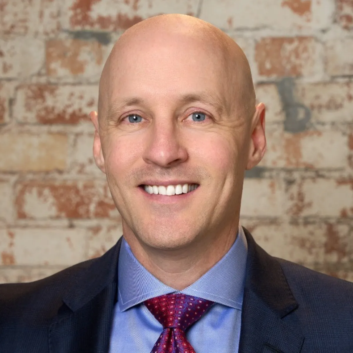 Tony Swantek in a suit, blue shirt, and red tie smiles in front of a textured brick wall.