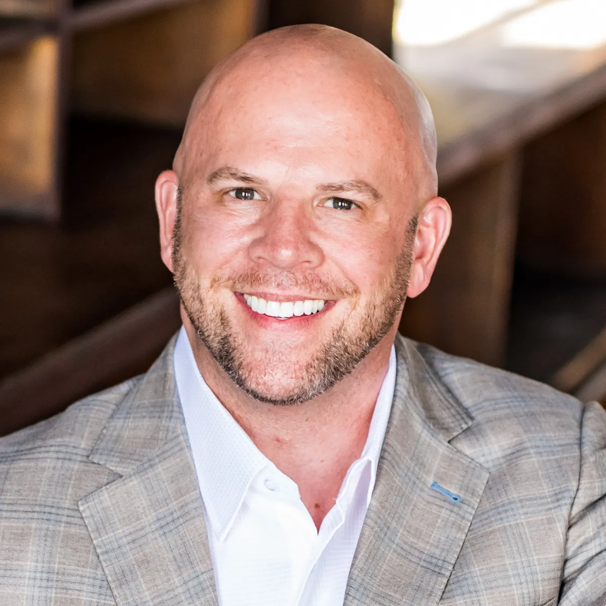 A smiling Darin Kidd with a trimmed beard, wearing a light plaid suit jacket and a white dress shirt, sits in front of a blurred wooden background.