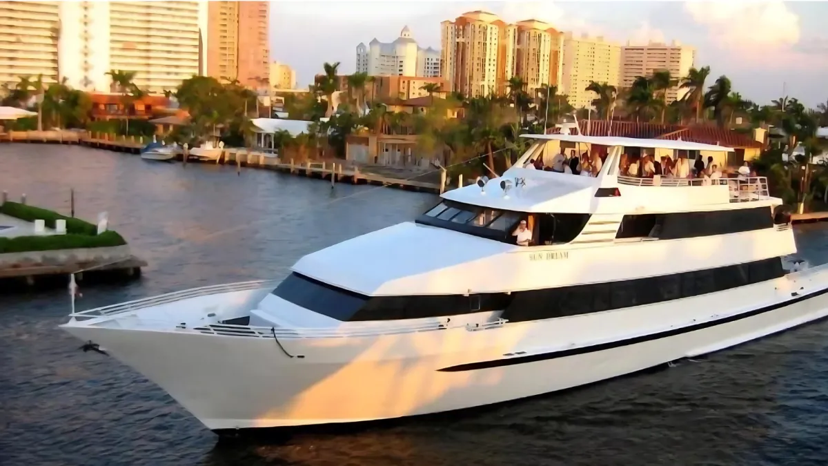 A large white yacht with multiple decks sails along a waterway, with people visible on the upper deck. Tall buildings, palm trees, and waterfront homes line the background under a partly cloudy sky at sunset.