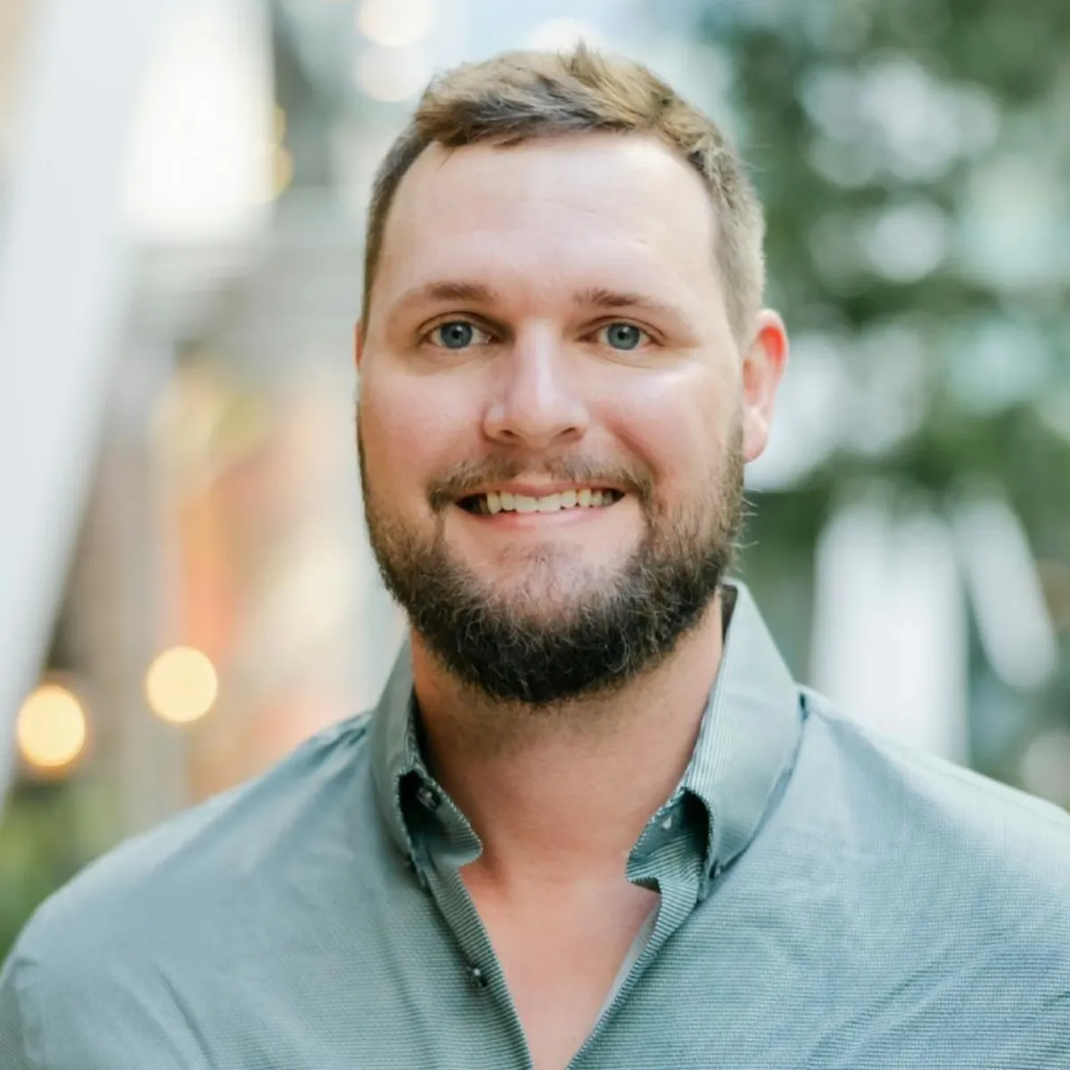 A smiling Darin Kidd with a trimmed beard, wearing a light plaid suit jacket and a white dress shirt, sits in front of a blurred wooden background.