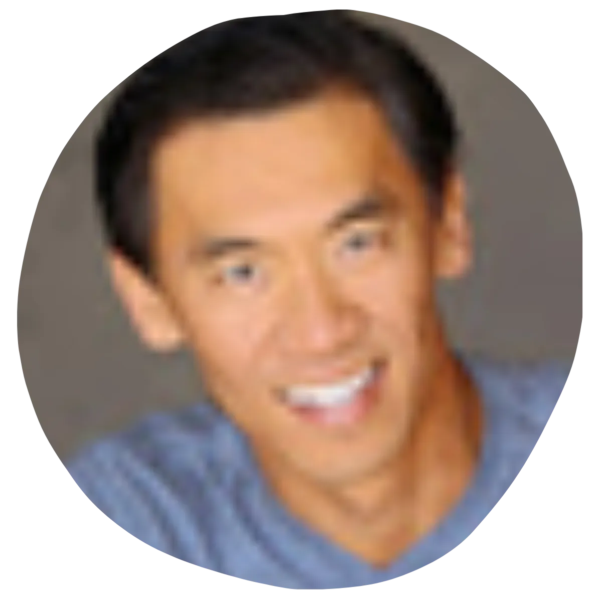 A man with short black and gray hair smiles warmly at the camera. He has a smooth skin complexion with visible laugh lines and is wearing a beige and brown striped shirt. The background is a solid dark brown color.