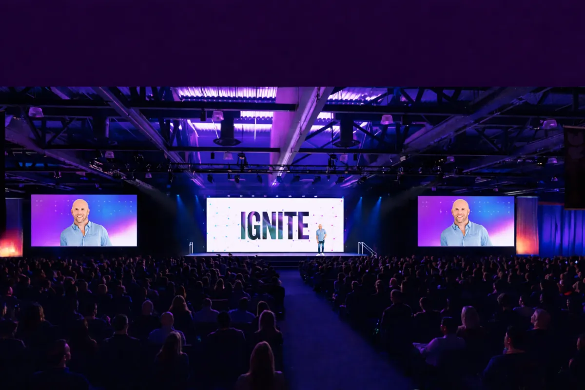 Large audience attending an R-Link Ignite live event, facing a main stage with a speaker and oversized digital screens under purple and blue lighting in a modern conference venue.