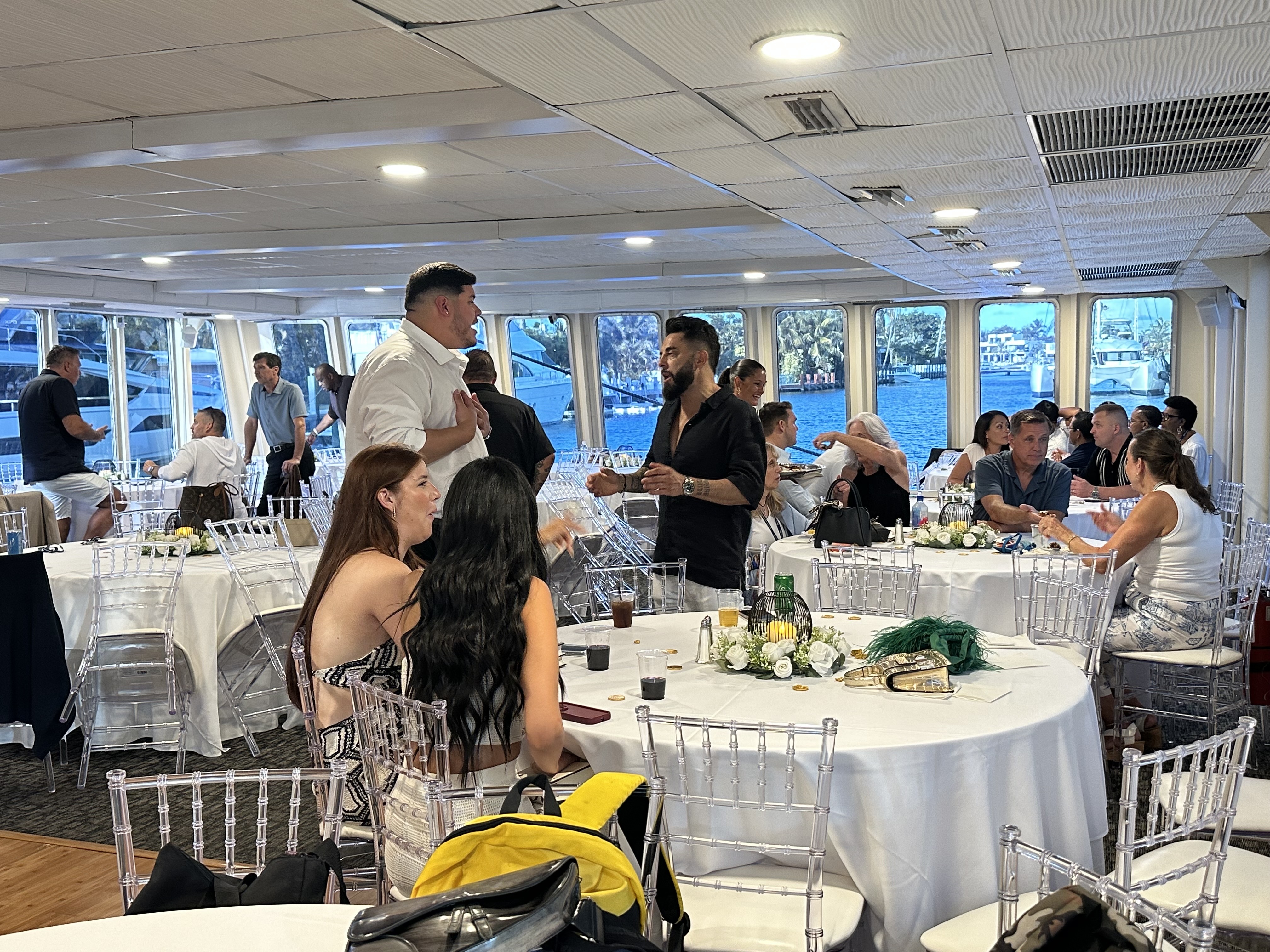Attendees gathered at an R-Link live event aboard a waterfront yacht, seated at round tables and networking inside a bright, modern venue with large windows overlooking the water and docked boats.