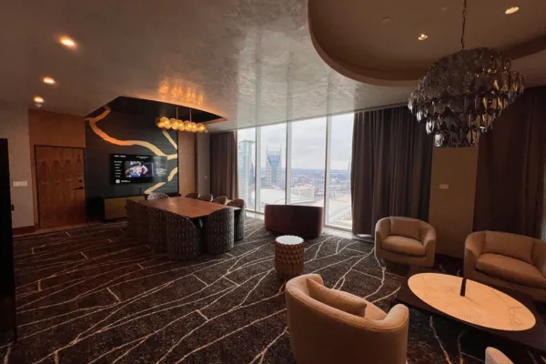 Renovated Broadway Suite living room at the Westin Nashville with panoramic floor-to-ceiling windows showcasing the Nashville skyline including the AT&T Batman Building, a large wood conference table, sculptural lounge chairs, a bold gold-veined black accent wall with flat-screen TV, dark patterned carpet, and a dramatic smoked-crystal chandelier