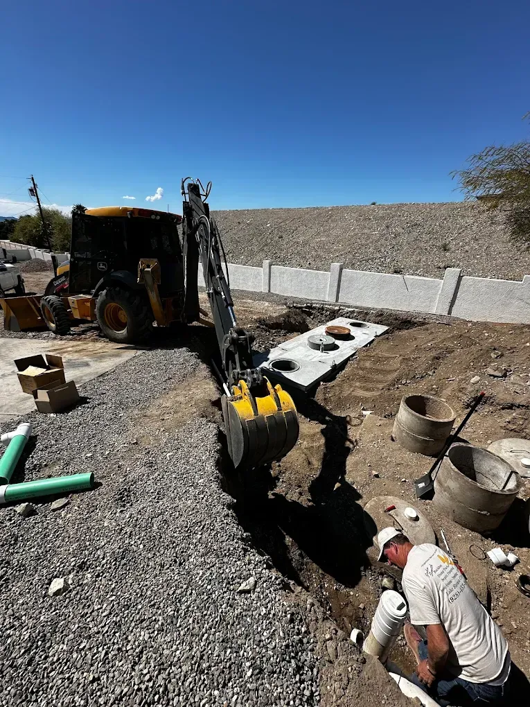 A diverse team of three construction professionals in safety vests and hard hats reviewing blueprints at a dusty job site, with heavy equipment in the background, under bright midday sun. The image is candid, documentary-style, and emphasizes teamwork and safety.