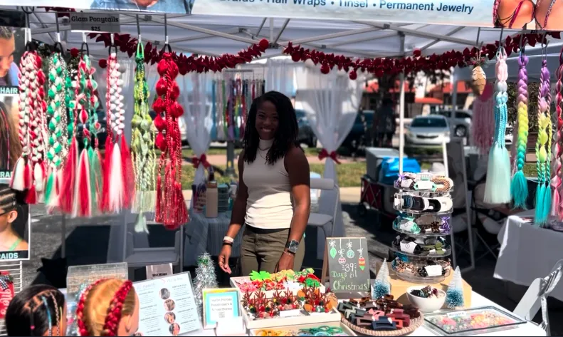 Woman standing at a vibrant market stall showcasing colorful hair accessories, including braids and hair wraps, with festive decorations and promotional signs.