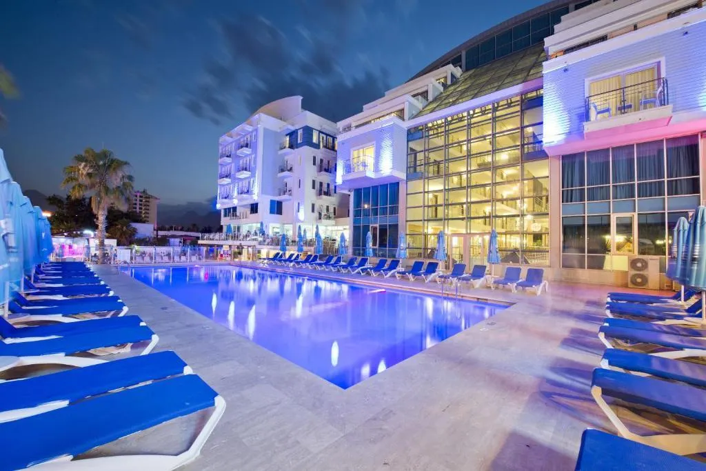 Piscine illuminée de nuit dans l'hôtel Sealife Family Resort  entourée de chaises longues bleues et d'une vue sur la façade vitrée du bâtiment. L'atmosphère est calme et élégante.