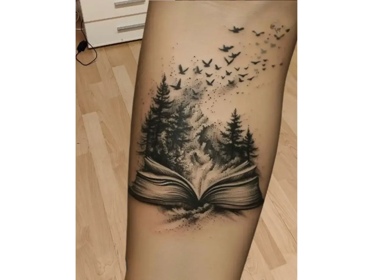 Tatouage réaliste en noir et gris représentant un livre ouvert d’où s’élèvent une forêt de pins et des oiseaux en vol – Magic Tattoo Ink