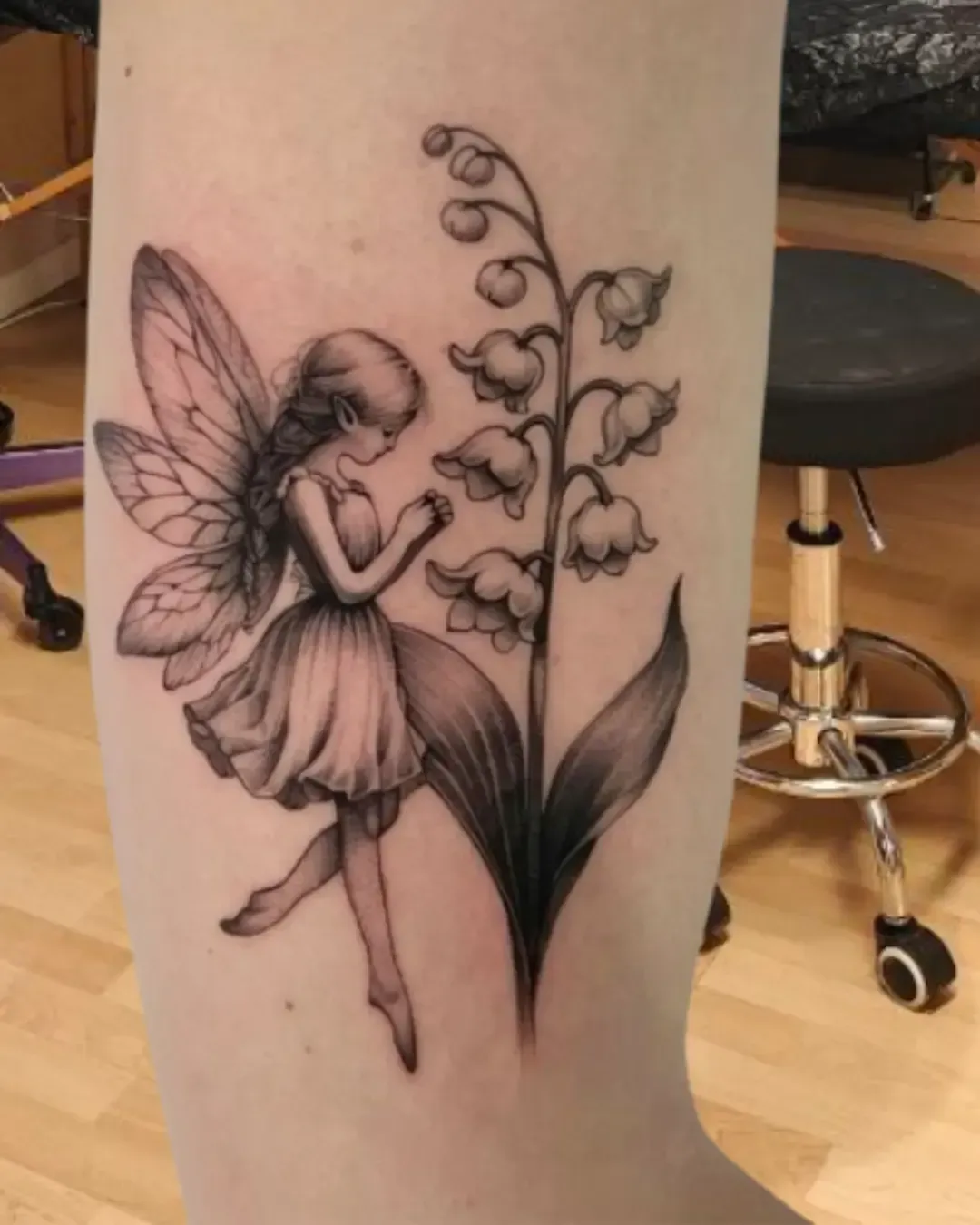 Tatouage poétique d’une fée délicate en noir et gris, agenouillée devant un brin de muguet en fleurs. Ce dessin mêle la magie des créatures féeriques à la symbolique du muguet, porte-bonheur et messager de joie. Un motif raffiné et onirique, parfait pour les amateurs d’univers enchanteurs et de nature. ✨🌿 Réalisé avec finesse par Magic Tattoo Ink à Fribourg.