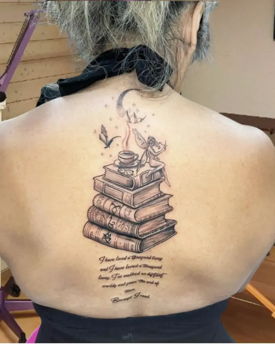 Tatouage poétique dans le dos représentant une pile de livres anciens, surmontée d’une fée assise près d’une tasse fumante, avec une lune et des étoiles en arrière-plan. En dessous, une citation inspirante rappelle la puissance de la lecture et des mondes imaginaires. Un chef-d'œuvre symbolique signé Magic Ink à Fribourg.