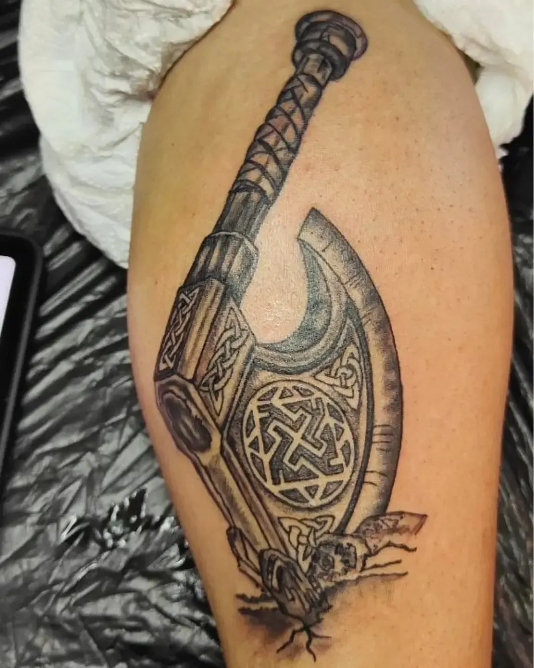 Tatouage d’une hache viking ornée de motifs celtiques, plantée dans le sol avec effet de fissures, symbole de force, courage et héritage nordique.