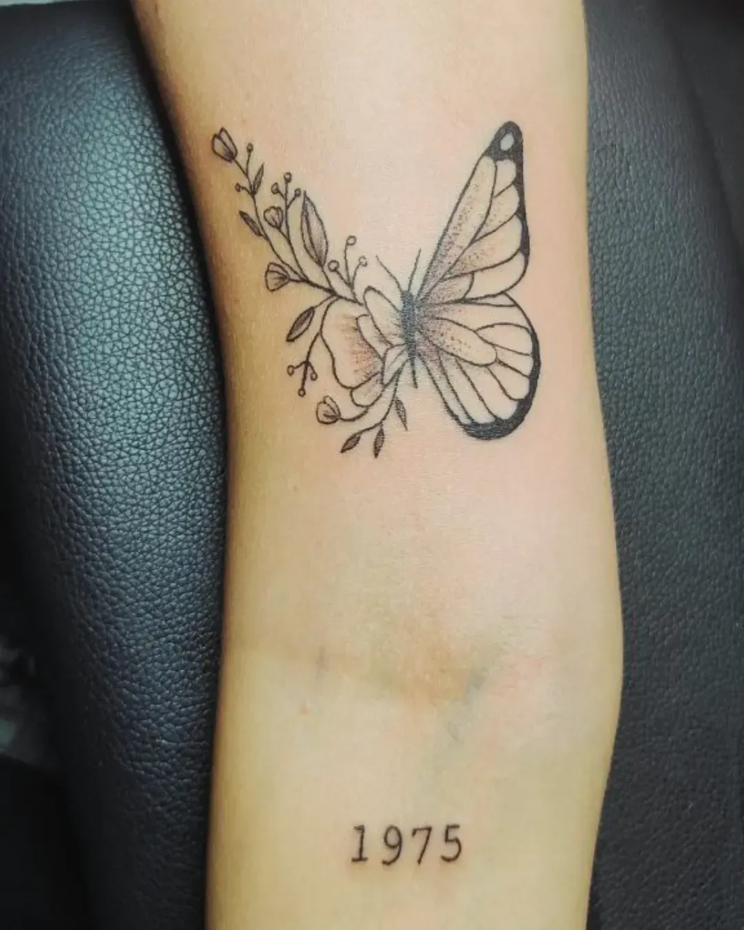 Tatouage d’un papillon délicatement fusionné avec des fleurs et des branchages fins, accompagné en dessous de l’inscription « 1975 », symbole personnel ou commémoratif.