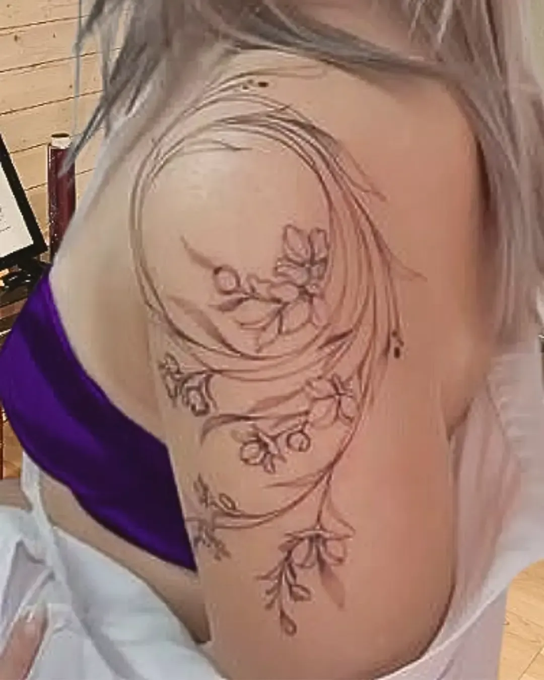Tatouage floral délicat sur l’épaule représentant un cercle artistique aux lignes fluides, entourant des branches fleuries aux pétales fins et élégants. Ce motif mêle harmonie et légèreté, rappelant la nature et le cycle de la vie. Une création raffinée de Magic Tattoo Ink à Fribourg, idéale pour sublimer le haut du bras avec poésie et féminité.