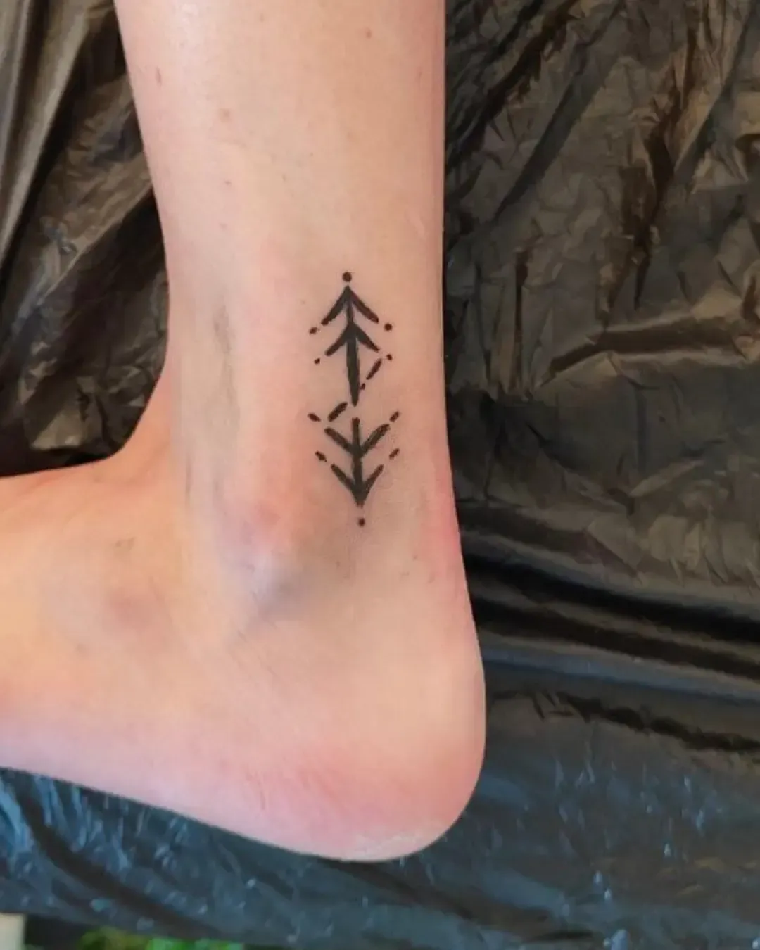 Tatouage minimaliste près de la cheville représentant deux flèches géométriques pointant vers le haut, accentuées de petits points décoratifs.