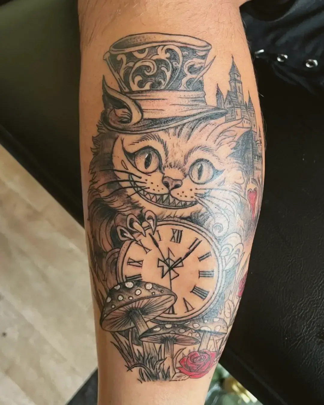 Tatouage artistique du Chat du Cheshire avec chapeau haut-de-forme, horloge, champignons et château – style Alice au Pays des Merveilles, réalisé par Magic Ink à Fribourg.