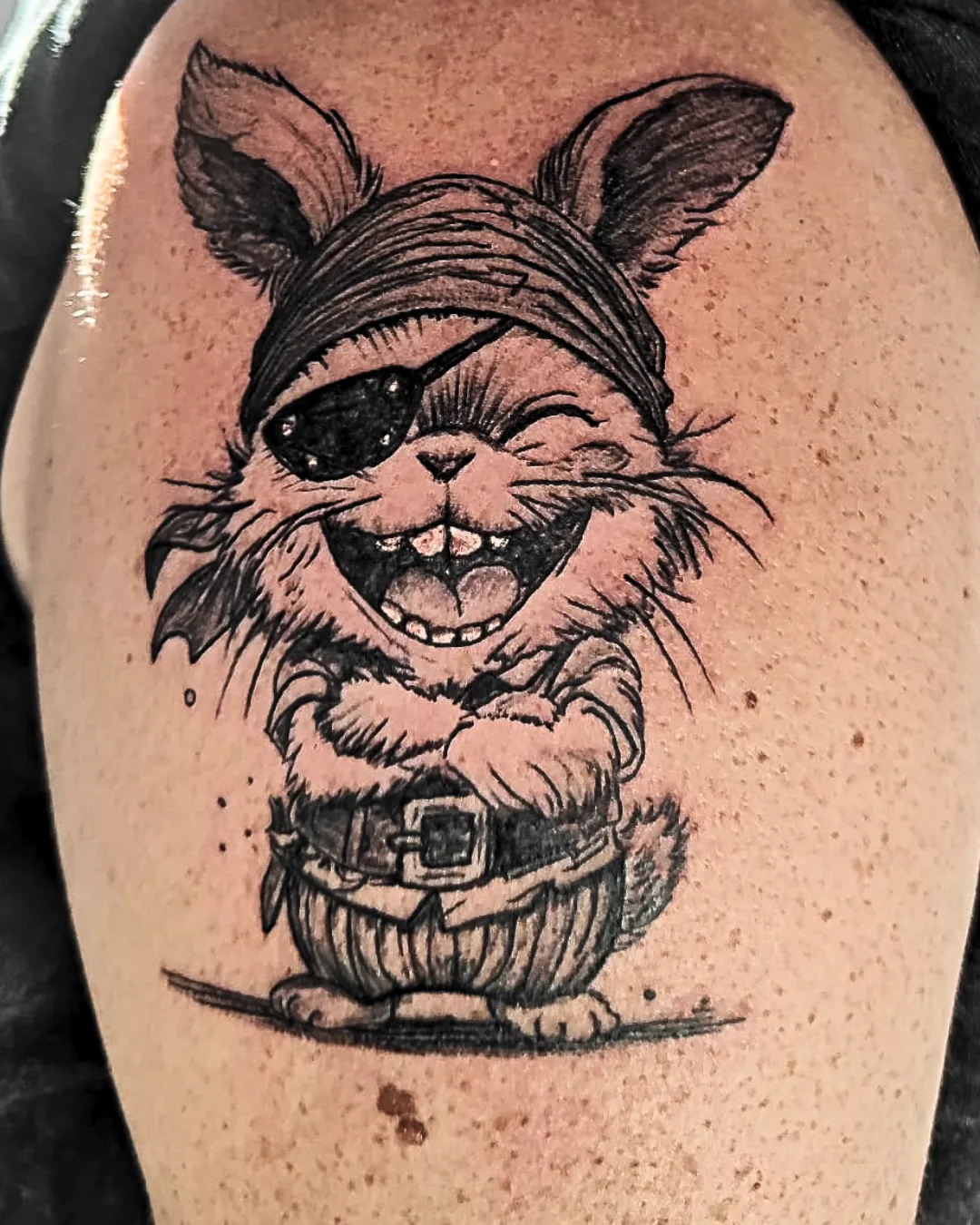 Tatouage fun et expressif d’un lapin pirate espiègle. Toujours avec son bandana et son cache-œil, mais cette fois-ci il arbore un grand sourire malicieux et un clin d’œil complice. Les bras croisés et l’attitude détendue renforcent son côté joueur et charismatique. Réalisé en noir et gris, ce dessin mêle humour et caractère, parfait pour ceux qui aiment afficher leur originalité avec légèreté.