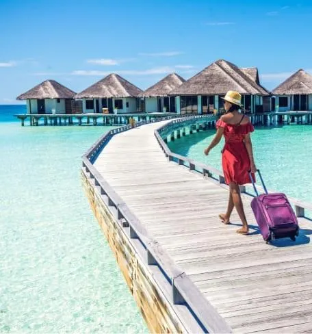 Vue d'un bungalow sur pilotis aux Maldives, avec une femme marchant sur un ponton en bois, portant une valise, dans un cadre paradisiaque aux eaux cristallines.