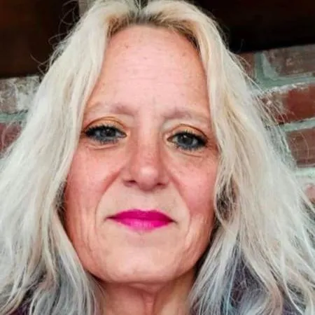 Woman with long, light-colored hair and bright pink lipstick, smiling against a brick background, representing the friendly and approachable team at Wow Wash 360.